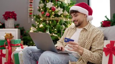 Şapkalı genç adam Noel hediyeleri, dizüstü bilgisayar ve kredi kartıyla evdeki kanepede oturuyor.