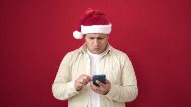 Noel Baba şapkalı adam cep telefonu kullanıyor ve kırmızı arka planda Noel 'i kutluyor.