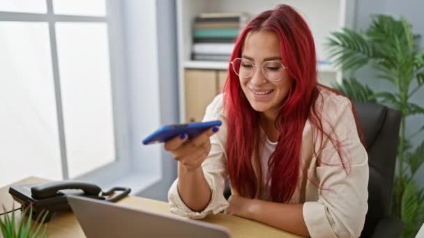 スマートフォンで魅力的な音声メッセージを送る若い女性労働者 オフィスの寿命を揺るがすスマイルビジネスに精通した赤毛 — ストック動画