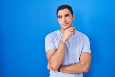 Young hispanic man standing over blue background with hand on chin thinking about question, pensive expression. smiling with thoughtful face. doubt concept. 