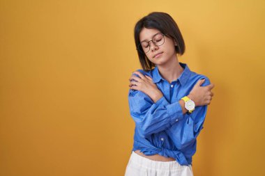 Sarı arka planda duran genç kız kendine sarılıyor mutlu ve pozitif, kendinden emin gülümsüyor. Sevgi ve özgüven. 