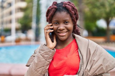 Afro-Amerikalı kadın gülümsüyor. Parktaki akıllı telefondan konuşuyor.