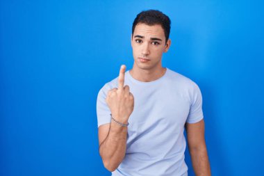 Young hispanic man standing over blue background showing middle finger, impolite and rude fuck off expression 