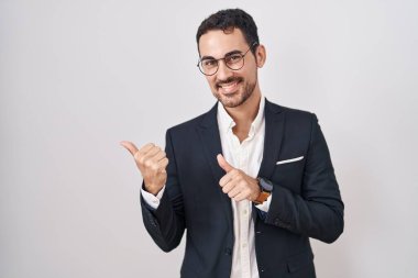 Beyaz arka planda duran yakışıklı İspanyol bir adam. Arka tarafı işaret ediyor. El ve baş parmakları havada, kendinden emin gülümsüyor. 