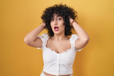 Sarı arka planda duran kıvırcık saçlı genç esmer kadın çıldırmış ve elleri başının üstünde korkmuş, ağzı açık şoktan korkmuş ve şaşırmış. 