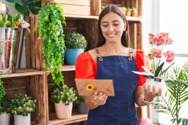 Young beautiful hispanic woman florist holding envelope letter and plant at florist