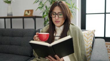 Young beautiful hispanic woman reading book and drinking coffee sitting on sofa at home