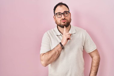 Artı, pembe arka planda duran sakallı, çenesinde parmak olan ve kafasını kaldırıp merak eden Latin kökenli bir adam. 