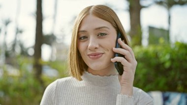 Genç sarışın kadın gülümsüyor. Parkta akıllı telefondan konuşuyor.