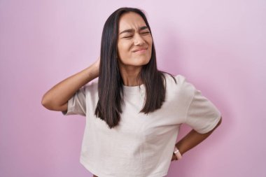 Young hispanic woman standing over pink background suffering of neck ache injury, touching neck with hand, muscular pain 