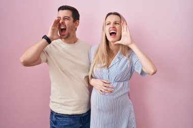 Genç bir çift pembe arka planda durup el ele verip bağırarak ve yüksek sesle çığlık atarak bebek bekliyor. iletişim konsepti. 