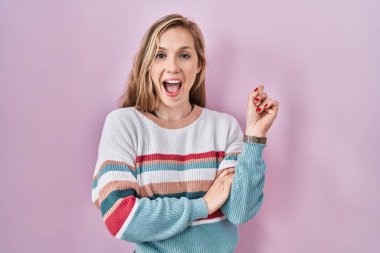 Pembe arka planda duran genç sarışın kadın yüzünde büyük bir gülümsemeyle, kameraya el ile işaret ederek bakıyor.. 