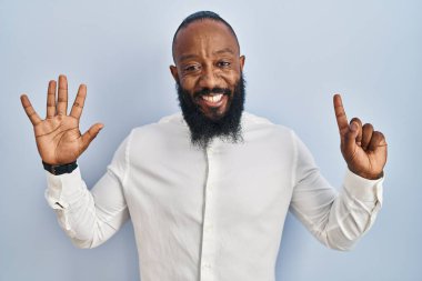 Mavi arka planda dikilen Afrikalı Amerikalı adam altı numaralı parmaklarını gösterip işaret ederken kendinden emin ve mutlu gülümsüyor.. 