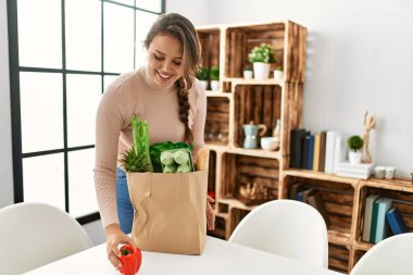 Genç, güzel, İspanyol bir kadın gülümsüyor. Elinde alışveriş torbası var.