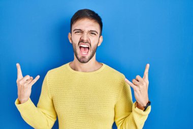 Hispanic man standing over blue background shouting with crazy expression doing rock symbol with hands up. music star. heavy music concept. 