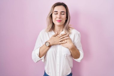 Pembe arka planda duran genç ve güzel bir kadın elleri göğsünde, gözleri kapalı ve yüzünde minnet dolu bir jest ile gülümsüyor. sağlık kavramı. 