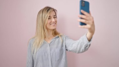 Genç sarışın kadın kendinden emin gülümsüyor. Akıllı telefonun yanında özçekim yapıyor. İzole edilmiş pembe arka planda.