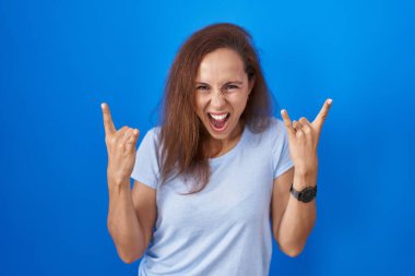 Mavi arka planda duran esmer kadın çılgın bir ifadeyle el kaldırarak rock sembolü yapıyor. Müzik yıldızı. ağır müzik konsepti. 