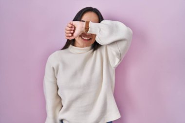 Pembe arka planda duran genç Güney Asyalı kadın kollarıyla gözlerini kapayarak neşeli ve komik gülümsüyor. kör kavram. 