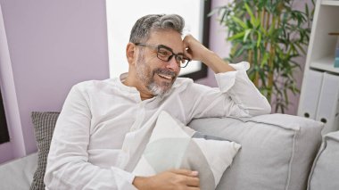 Neşeli, gri saçlı İspanyol adam evdeki oturma odasındaki kanepede mutlu ve rahat bir şekilde oturur, güvenle gülümser..