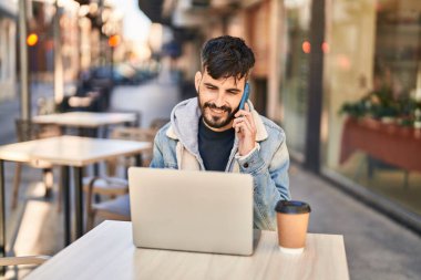 Young hispanic man using laptop talking on smartphone sitting on table at coffee shop terrace
