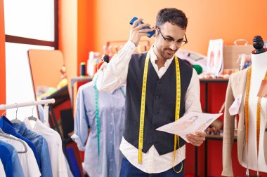Young hispanic man tailor listening voice message by smartphone looking clothing design at atelier