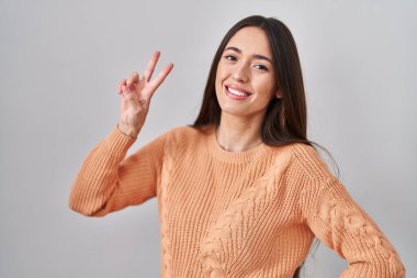 Beyaz arka planda duran esmer kadın kameraya bakıp zafer işareti yapıyor. İki numara.. 