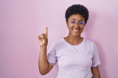 Young african american woman standing over pink background showing and pointing up with finger number one while smiling confident and happy. 