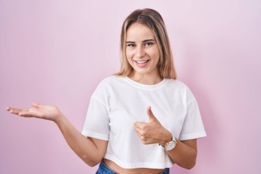 Pembe arka planda duran genç sarışın kadın avuç içini gösteriyor ve baş parmaklarıyla iyi bir hareket yapıyor, mutlu ve neşeli gülümsüyor. 