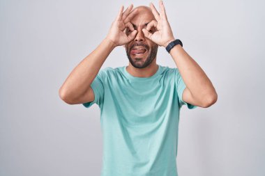 Orta yaşlı kel bir adam beyaz arka planda dikiliyor. Dürbün gibi jest yapıyor. Dürbün dilini çıkarıyor, gözler parmaklarından bakıyor. çılgın ifade. 