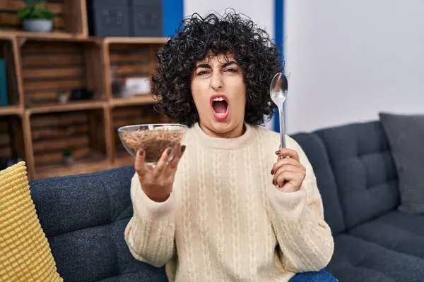 Young brunette woman with curly hair eating healthy whole grain cereals with spoon angry and mad screaming frustrated and furious, shouting with anger. rage and aggressive concept.