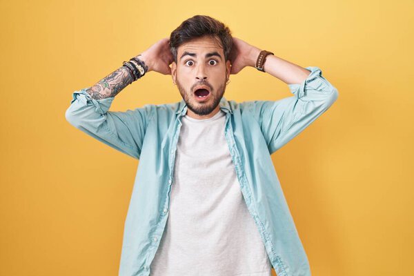 Young hispanic man with tattoos standing over yellow background crazy and scared with hands on head, afraid and surprised of shock with open mouth 