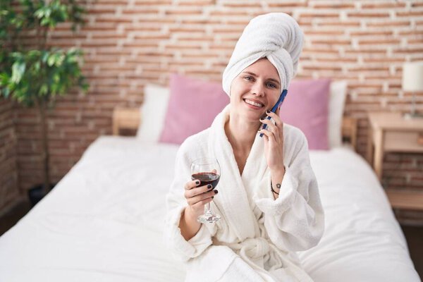 Молодая блондинка разговаривает по смартфону и пьет вино в спальне.