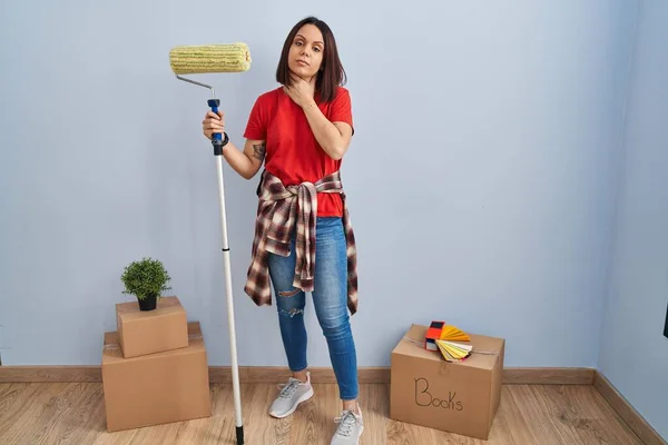 Young hispanic woman painting home walls with paint roller touching painful neck, sore throat for flu, clod and infection