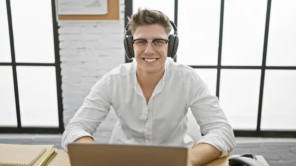 Smiling young caucasian business worker enjoying his job, confidently managing work online on laptop, with headphones on his desk at the office.