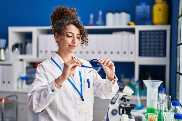 Young hispanic woman scientist smiling confident holding security glasses at laboratory