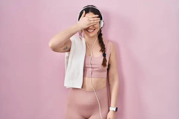 Young brunette woman wearing sportswear and headphones smiling and laughing with hand on face covering eyes for surprise. blind concept.