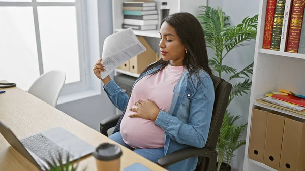 年轻的怀孕女商人在炎热的办公室里与不适感作斗争 一边把文件当作手扇一边埋头苦干 — 图库照片