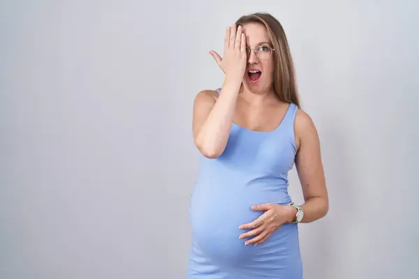 年轻的孕妇站在白底之上 用一只手捂住一只眼睛 脸上带着自信的微笑和惊讶的情绪 — 图库照片