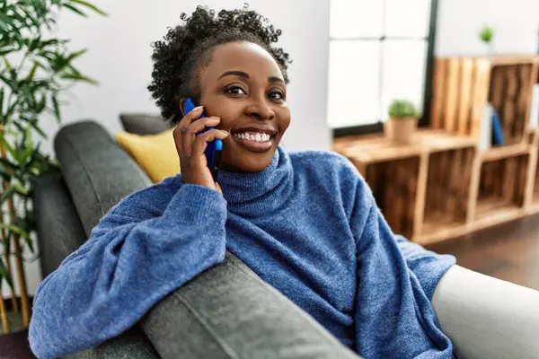 アフリカ系アメリカ人の女性が自宅でソファに座っているスマートフォンで話しています — ストック写真