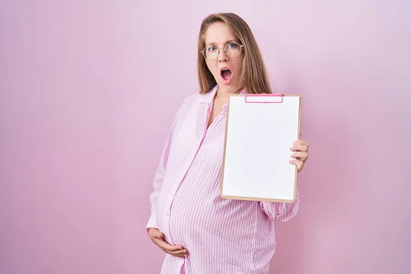 ショックの顔にクリップボードを保持している若い妊婦 懐疑的で皮肉を見て 開いた口で驚いた — ストック写真