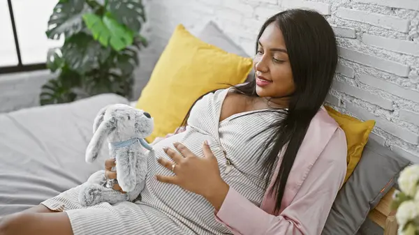 Young, expecting mother tenderly touches her pregnant belly, lying relaxed in bed with a rabbit toy in a cozy bedroom setting, radiantly expressing her joyous anticipation of motherhood.