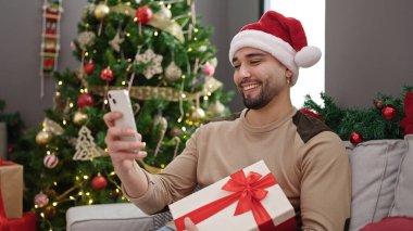 Genç Arap adam elinde akıllı telefon hediyesiyle evde Noel ağacının yanında oturuyor.