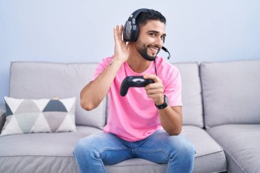 İspanyol asıllı genç adam video oyunu oynuyor kanepede oturmuş, sırıtarak dedikodu ve dedikoduları dinliyor. Sağırlık kavramı. 