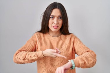 Genç esmer kadın beyaz arka planda dikiliyor. Aceleyle zamanı, sabırsızlığı ve gecikmeyi gösteriyor. 