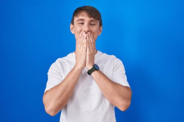 Mavi arka planda dikilen beyaz, sarışın adam kıkırdıyor, gülüyor, kıkırdıyor elleriyle ağzını kapatıyor, dedikodu yapıyor ve skandal konsepti yapıyor. 