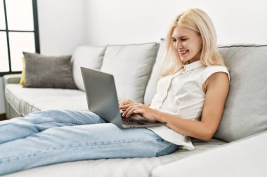 Evdeki kanepede dizüstü bilgisayar kullanan genç sarışın kadın.