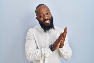 Afro-Amerikalı adam mavi arka planda ayakta alkışlıyor ve alkışlıyor mutlu ve neşeli, gururla gülümsüyor. 