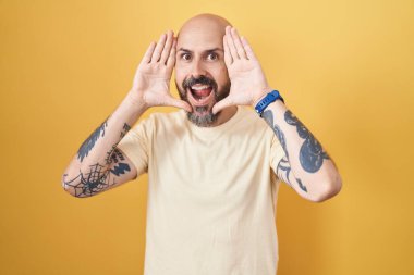 Hispanic man with tattoos standing over yellow background smiling cheerful playing peek a boo with hands showing face. surprised and exited 