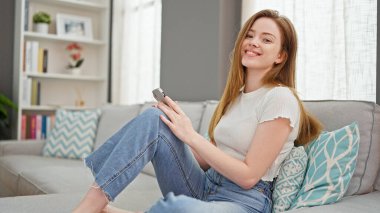 Evdeki kanepede oturan akıllı telefon kullanan genç sarışın kadın.